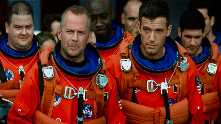 Willis e Affleck estão lado a lado, vestidos de astronautas, com macacões laranjas. Atrás deles há mais homens vestidos da mesma forma