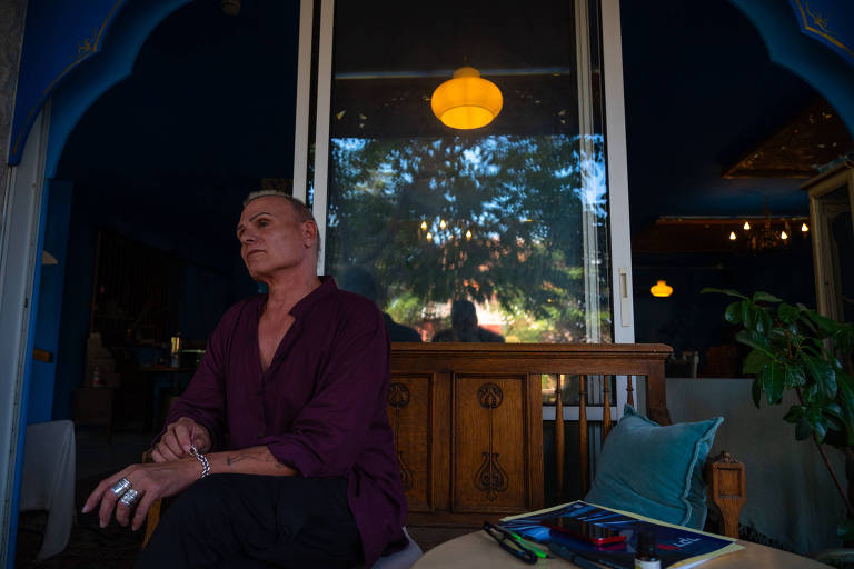homem branco, está sentado virado para a esquerda da foto, usa camisa roxa e calça preta, atrás dele um móvel de madeira e um lustre com luz amarela acima, por trás de um vidro. à direita do homem, uma mesa com uma revista e uma almofada azul em uma cadeira 