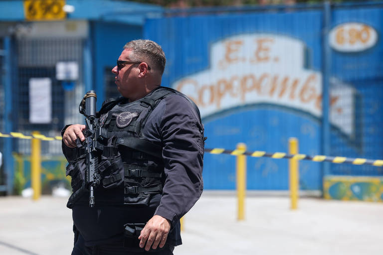 Há um policial vestido de preto e armado na frente do muro da escola, que é azul