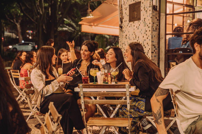 Pessoas são vistas sendas em mesa do lado de fora de um bar; elas estão sorrindo e conversando