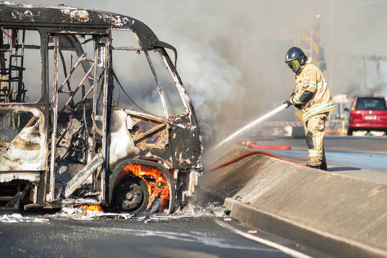 Bombeiro com mangueira joga água em carcaça de ônibus queimada e ainda com chamas