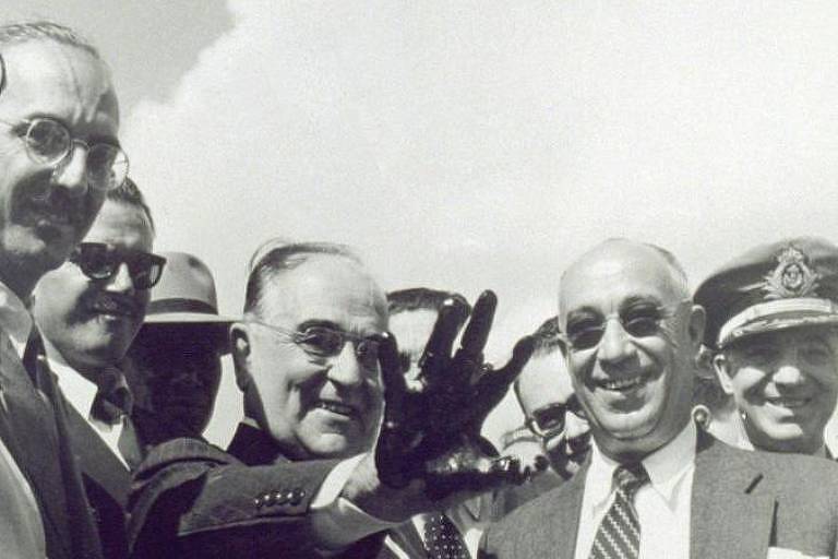 Getúlio Vargas sorri ao mostrar mão suja de petróleo, rodeado por vários homens engravatados

