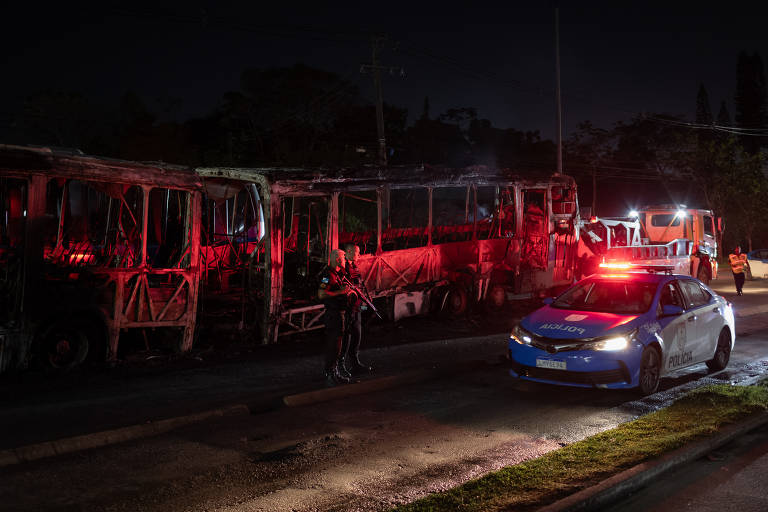 Carcaça de ônibus queimado. À frente, policiais militares e uma viatura