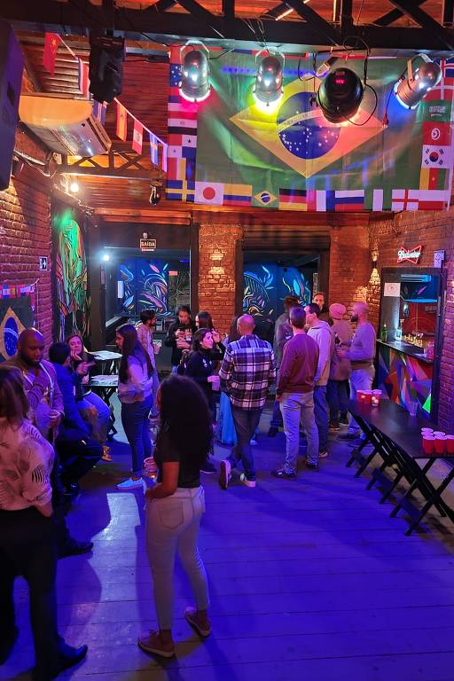 No Supra Bar Vila Madalena, participantes do projeto Multilingo Language Exchange circulam com adesivos colados na roupa correspondentes aos idiomas em que são fluentes ou que gostariam de treinar; o ambiente é decorado com bandeiras de diferentes países