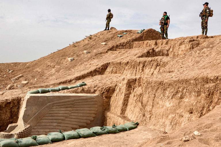 A imagem mostra um terreno árido e montanhoso， onde três soldados estão posicionados em um ponto elevado. Eles estão vestidos com uniformes militares e observam a área ao redor. Na parte inferior da imagem， há uma estrutura de concreto com sacos de areia ao redor， sugerindo uma fortificação ou abrigo. O céu está claro， e a paisagem é predominantemente marrom e seca.