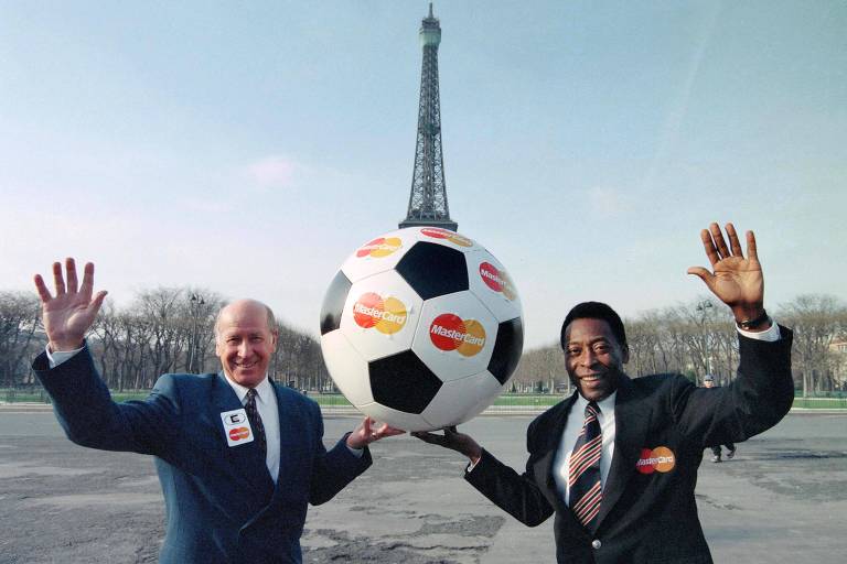 Bobby Charlton e Pelé em foto em Paris com a Torre Eiffel atrás deles; os dois usam terno e gravata e seguram, juntos, uma bola de futebol gigante
