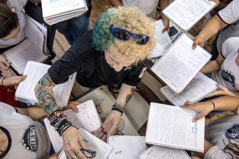 Mulher branca, de cabelo loiro, curto e encaracolado, com tatuagem no braço direito, carimba o caderno de um de seus alunos; outros fazem um círculo de cadernos ao redor dela