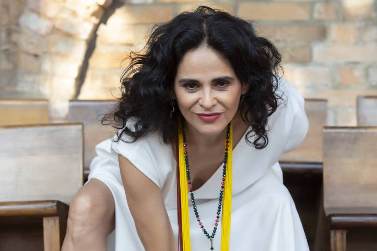 Em foto colorida, a cantora Mariana de Moraes posa para a câmera com vestido branco e vários colares coloridos