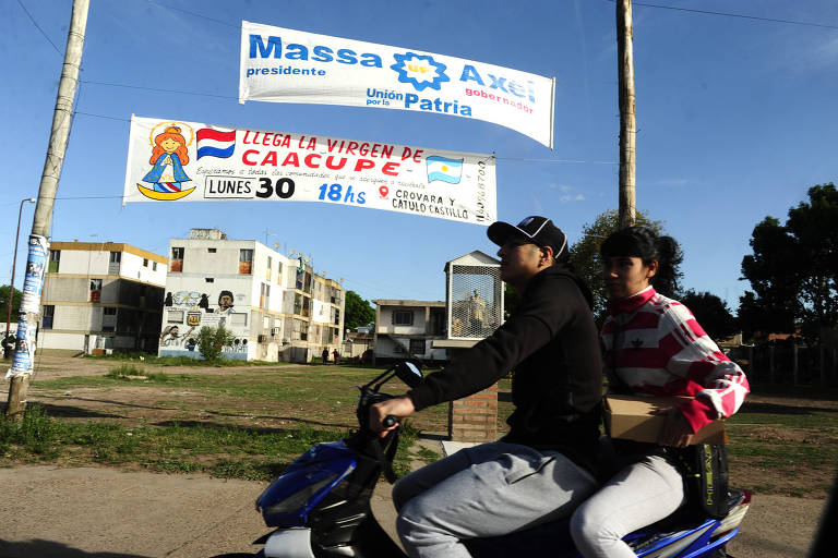 Homem e mulher passam em moto em frente a faixa pendurada em postes que diz "Massa"