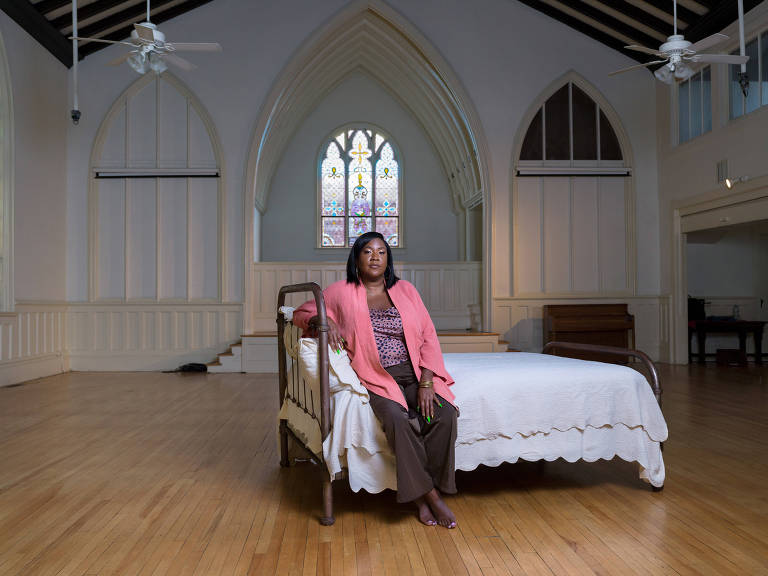 Fotografia colorida mostra Tricia Hersey, uma mulher negra que veste uma calça marrom e uma blusa e um casaquinhos, ambos da cor rosa; ela está sentada em uma cama com lençois brancos dentro de uma catedral vazia