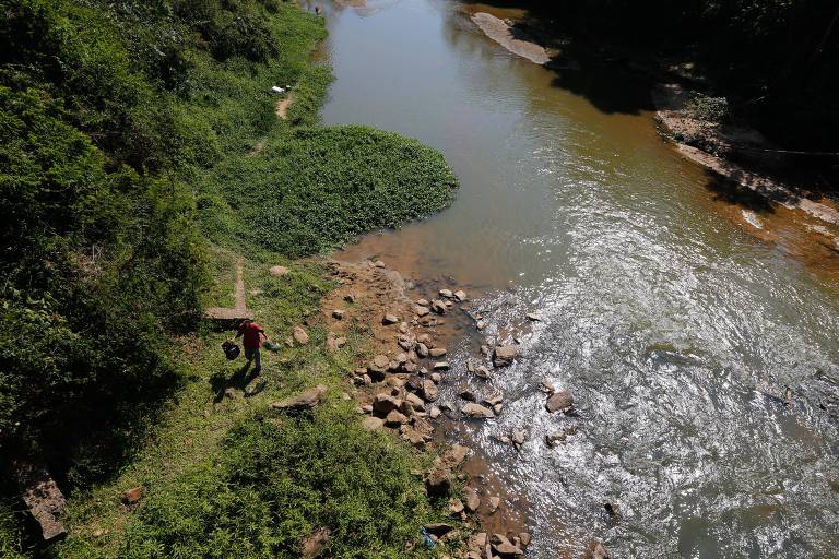Vista aérea do rio; em uma das margens, uma pessoa carrega um balde