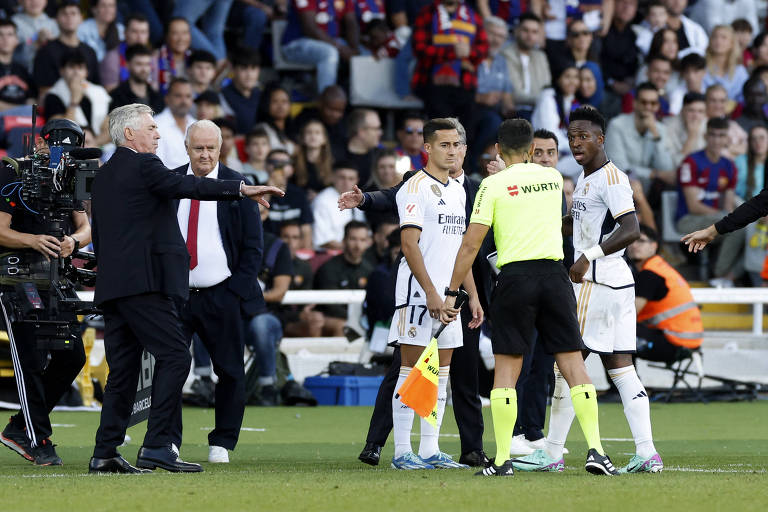 Vini Jr., à direita, discute com o bandeirinha. À esquerda, o técnico Ancelotti tem o braço esticado, chamando o jogador para apressá-lo