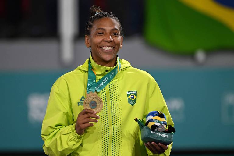 Rafaela, uma mulher negra, sorri e mostra com a mão direita a medalha de ouro, que leva pendurada no pescoço, no pódio. na mão esquerda ela tem um bichinho de pelúcia do torneio