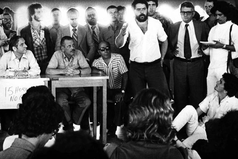 Em imagem de 1979, Lula transmite instruções recebidas da polícia para desocupação de prédio em intervenção do sindicato, em São Bernardo do Campo (SP); De pé, além de Lula, os deputados Antonio Resk e Alberto Goldman. Na mesa, o prefeito Tito Costa e os delegados de polícia Nivaldo Leme e Deodato Araújo
