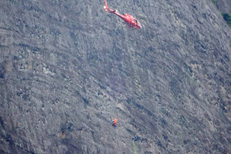 Na foto, helicóptero vermelho dos bombeiros resgata homem preso em rocha