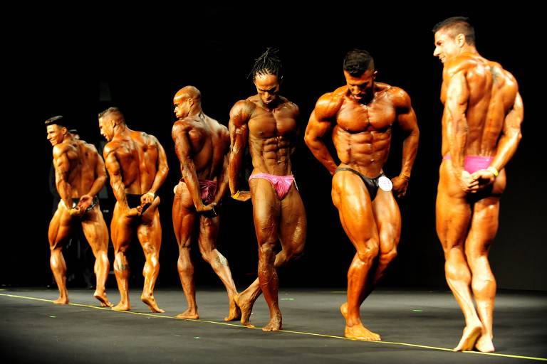 6 homens musculoso e bronzeados fazem poses em um palco, na frente de um fundo preto. Todos vestem sunga.