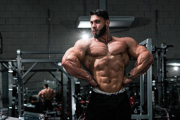 Ramon é um homem branco e musculoso. Ele está em uma academia de musculação e faz pose para a câmera, estufando o peitoral. Ele está sem camisa e veste uma bermuda preta.