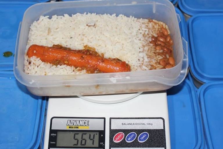 Marmita com arroz, feijão e uma salsicha em cima de uma balança, marcando o peso de 569 gramas
