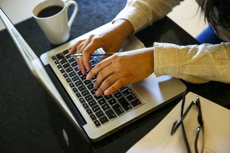 Foto foca em laptop sobre uma mesa. Mostra também mãos de uma pessoa ao digitar no teclado. Cada lado do computador tem um objeto, sendo um par de óculos e uma xícara de café