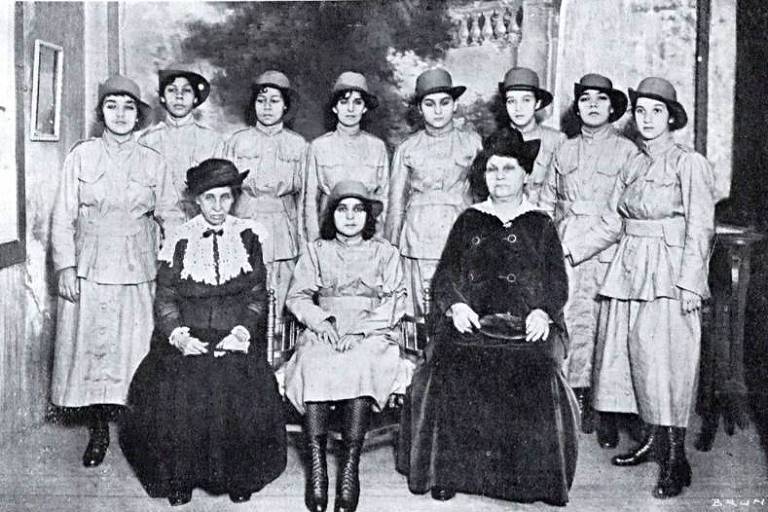 a foto, em preto e branco, dá destaque a duas mulheres que se vestem de preto, Izabel Dillon e Leolinda Daltro, e diversas jovens que a rodeiam num uniforme de cor clara que lembra uma vestimenta militar.