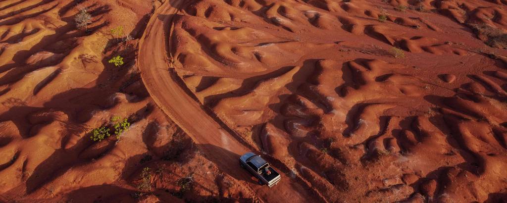 Vista de drone de carro andando em estrada em meio a um deserto de terra vermelha, com relevo cheio de 'dobras'
