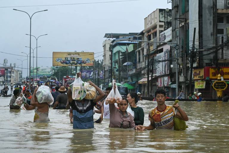 Pessoas andam com água pela cintura em uma rua, carregando com os braços erguidos sacolas de supermercado