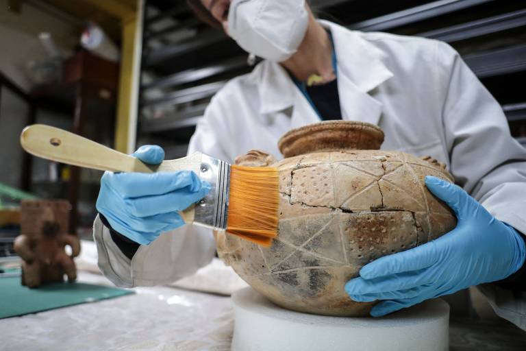Foto mostra restauradora passando um pincel em um recipiente de barro fragmentado. Ela usa luvas de látex azuis, jaleco e máscara