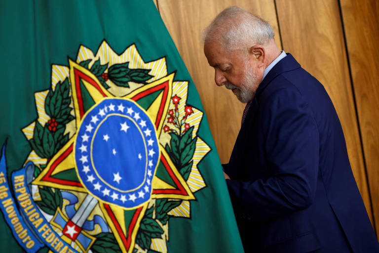 Lula de perfil, do quadril para cima. Ele olha para baixo e veste terno azul. No canto direito da imagem, há uma bandeira com o brasão da República