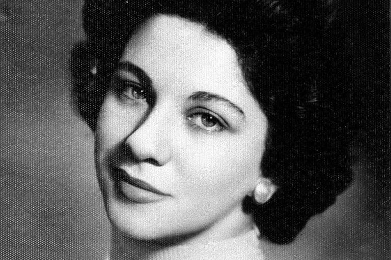 foto em preto e branco de mulher de cabelo preto e olhos claros, sorrindo com blusa branca de gola alta