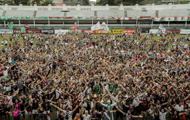 Uma multidão de pessoas está no gramado do estádio, com os braços abertos, festejando, voltados para a câmera.