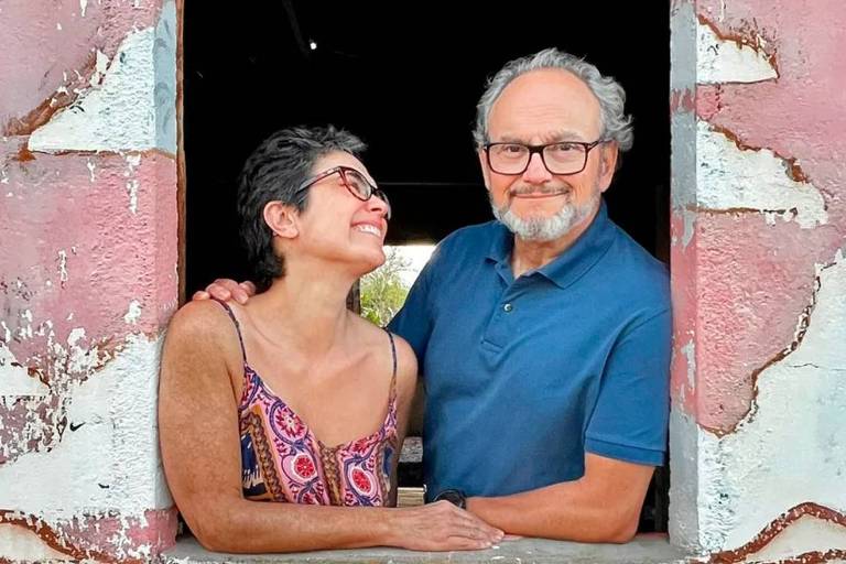 Em foto colorida, casal posa na janela de uma casa