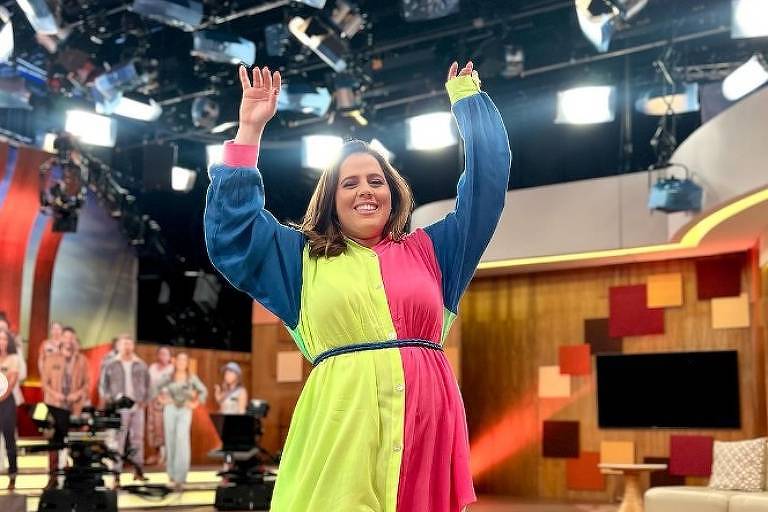Em foto colorida, mulher de vestido colorido levanto os braços e comemora o sucesso de um program de TV