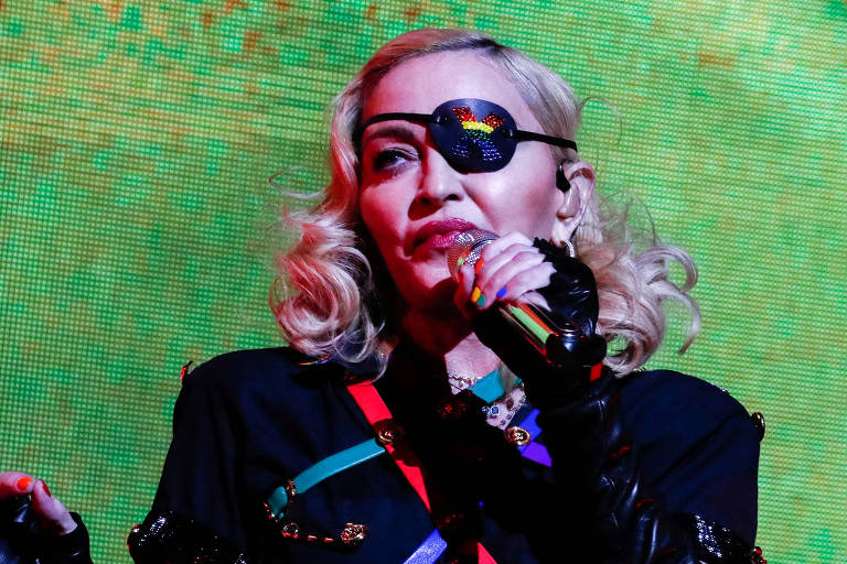 Cantora Madonna, uma mulher de pele branca e cabelos loiros, durante um show. Ela usa roupas pretas e um tapa-olho, Ao fundo, um telão verde.