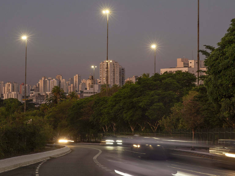 Imagem mostra postes de iluminação pública, sem fios, e reflexos dos faróis de carros na avenida
