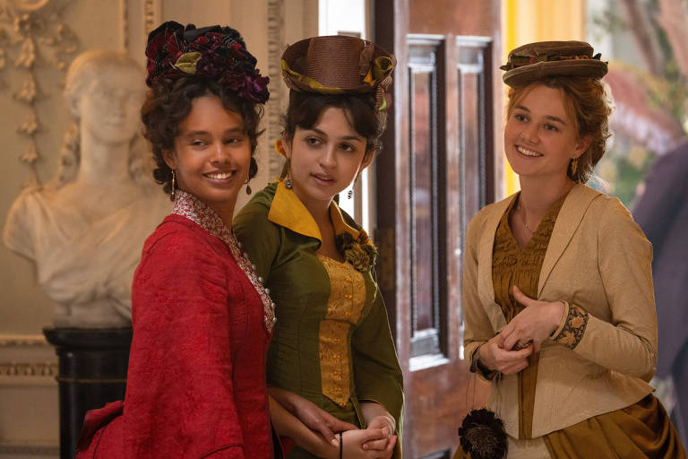 Três jovens mulheres em roupas dos anos 1870 conversam e sorriem em cena da série 