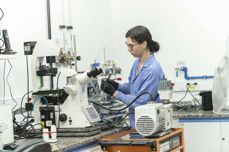 Mulher vestida com jaleco azul, luvas pretas e óculos opera um equipamento no laboratório da PUC-Rio