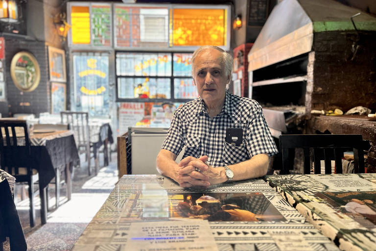 Um homem de camisa xadrez de mangas curtas está sentado, sozinho, à mesa de um restaurante; ao fundo, mesas vazias e uma grande churrasqueira