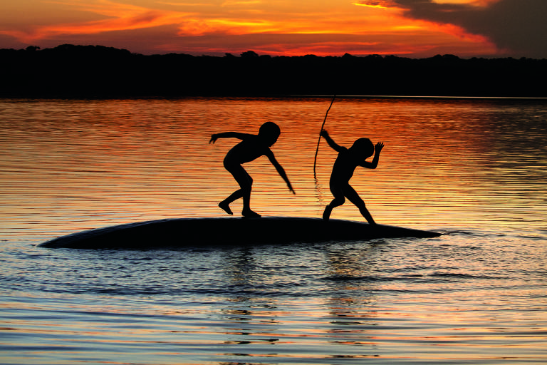 silhueta de duas crianças brincando em cima de canoa virada em rio