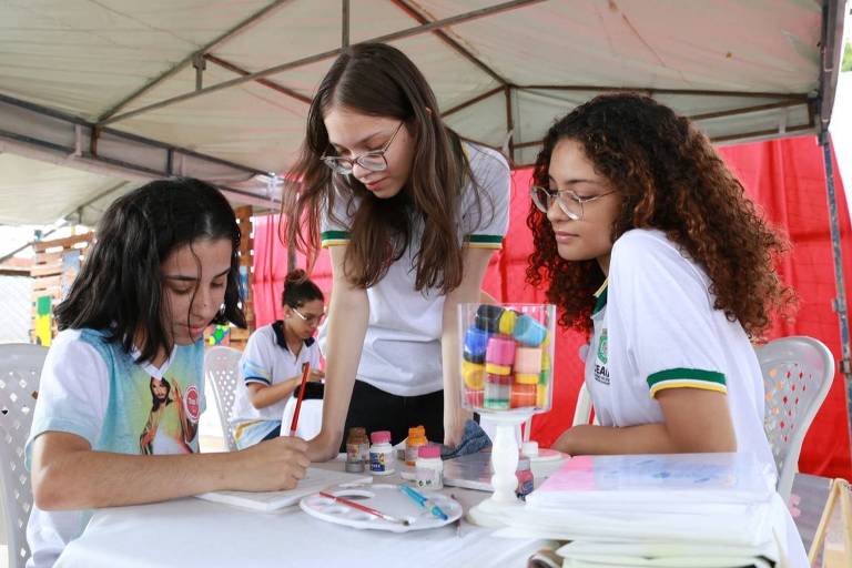 Imagem mostra três alunas vestindo uniforme da escola com pontes de tinta. Elas olham para um papel onde uma delas está fazendo um desenho. O uniforme é branco com litras verdes na manga.