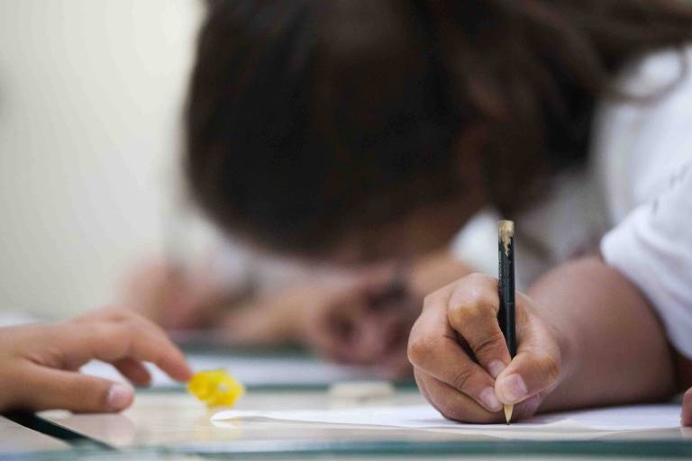 foto colorida com foco na mão de criança escrevendo com lapis em um papel