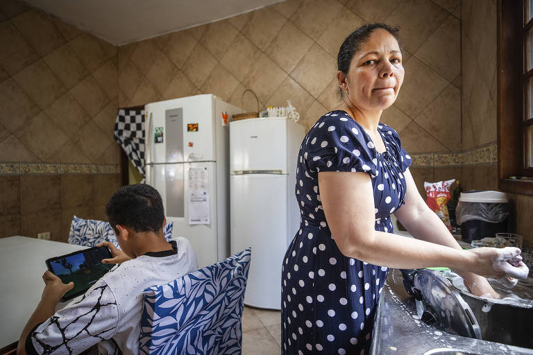 Mulher na cozinha lava louça, enquanto criança está sentada em mesa ao lado, no tablet