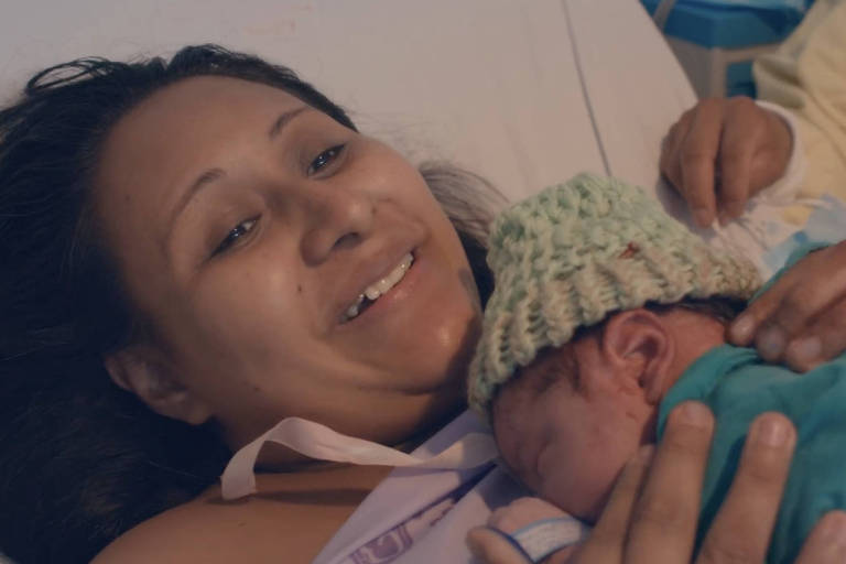 Uma mulher está deitada, sorrindo e segurando seu bebê recém-nascido sobre o corpo 