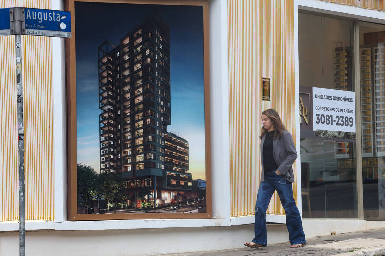 O homem tem cabelos longos, usa paletó cinza e calça jeans; a placa com o nome "Rua Augusta" está à frente de uma grande imagem de um edifício