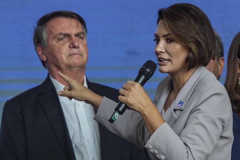 foto colorida com Michele à frente, com o microfone na mão e roupa cinza. Bolsonaro, homem idoso, está ao fundo sem sorriso 