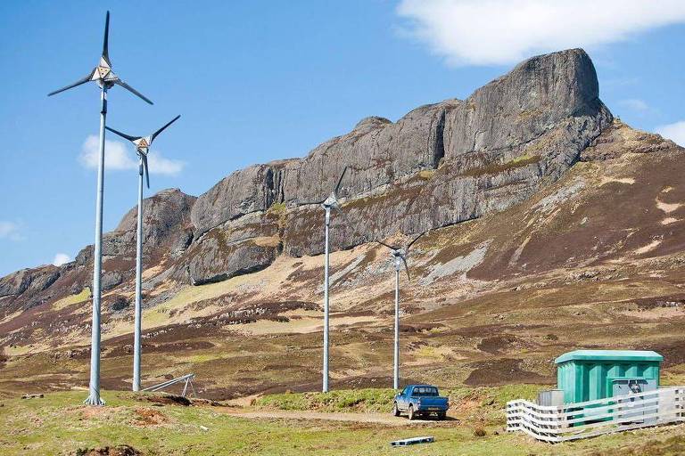 Cata-ventos gigantes de energia eólica em um campo ao lado de um morro; na planície há também uma camionete e uma pequena casa