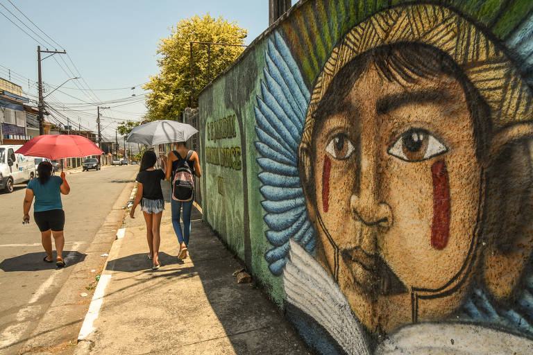 pessoas caminham sob o sol usando guarda-chuva para proteção; ao longo da calçada, muro com ilustração de pessoa indígena ocupa parte do lado direito da imagem 