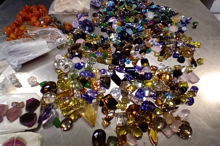Foto apresenta um conjunto de pedras preciosas de diversos tamanhos, como esmeraldas verdes, cristais de quartzo roxo e amarelo, colocados sobre uma mesa preta de forma aleatória