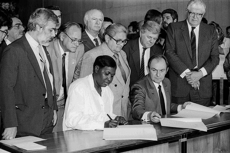 Benedita, mulher negra, está sentada e assina a Constituição; está ao lado de outro homem sentado e outros, em pé, estão atrás dela