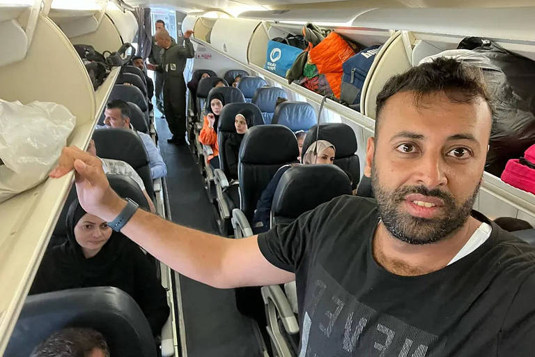 Homem de pele morena e barba, na faixa dos 35 anos, de pé dentro de um avião, aponta para outros passageiros nas poltronas.