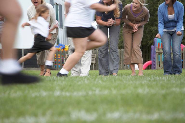 Crianças correndo em gramado, com mulheres no plano de fundo torcendo pelas crianças